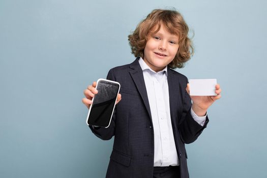 Self-confident Handsome positive happy boy with curly hair wearing suit holding phone and credit card and showing mobile screen at camera isolated over blue background looking at camera. Mockup