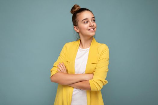 Photo shot of pretty positive happy smiling brunette little girl wearing stylish yellow jacket and white t-shirt standing isolated over blue background wall looking to the side. Empty space, copy space