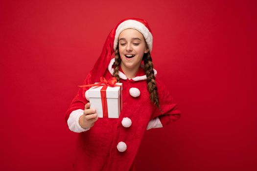 Shot of attractive happy smiling brunette girl isolated over red background wall wearing Santa Claus suit holding white gift box with red ribbon and looking at surprise.