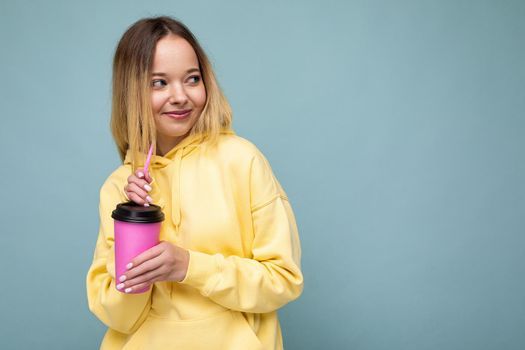 Beautiful young positive happy smiling blonde woman wearing yellow hoodie isolated over blue background holding paper cup for mockup drinking coffee and looking to the side. copy space