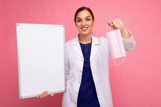 Woman doctor in a white medical coat holding blank board with copy space for text and protective mask isolated on background. Covid concept.