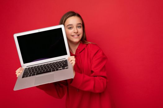 Close-up portrait photo of beautiful happy smiling girl with long hair wearing red hoodie holding computer laptop looking at camera isolated over red wall background. Cutout, copy space
