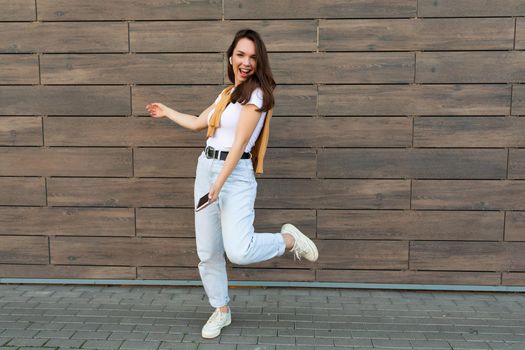 Full length body size photo of pretty brunet young happy woman wearing casual clothes and listening to music via wireless earphones standing in the street holding and using mobile phone looking at camera.Copy space