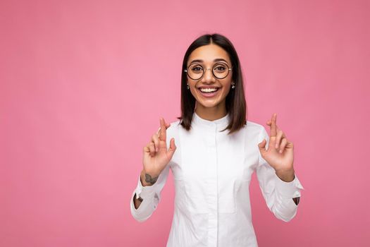 Portrait of young positive happy attractive brunette woman with sincere emotions wearing casual white shirt and optical glasses isolated over pink background with copy space and holding fingers crossed for good luck.