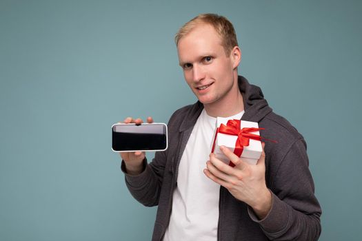 Shot of handsome positive smiling young blonde man isolated over blue background wall wearing casual stylish clothes holding white gift box with red ribbon and mobile phone with empty screen display for mockup looking at camera.