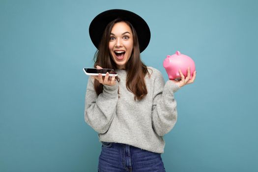 Portrait photo of happy positive sincere young charming winsome woman with dark hair wearing casual gray sweater and black hat isolated on blue background with copy space, holds pink pig money box and uses mobile phone communicating online.