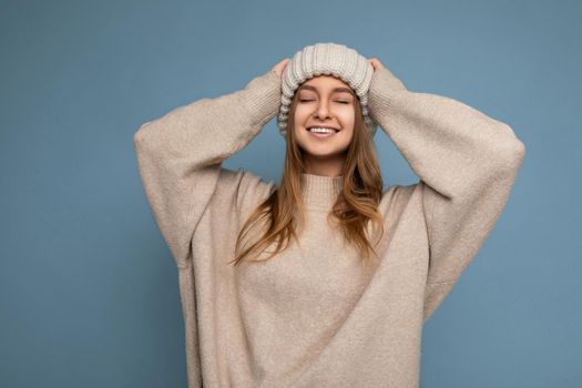 Shot of fascinating happy smiling adult dark blonde woman isolated over blue background wall wearing beige warm sweater and knitted beige hat enjoying with close eyes.