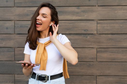 Beautiful brunet young happy woman wearing casual clothes and listening to music via wireless earphones standing in the street holding and using mobile phone.Copy space