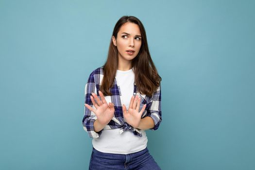 Young serious beautiful brunette woman with sincere emotions wearing trendy check shirt standing isolated on blue background with empty space and showing stop gesture saying no. Negative concept.