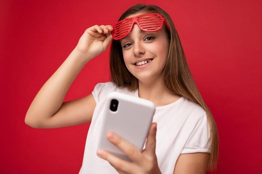 Photo shot of smiling attractive positive good looking teenager wearing casual stylish outfit poising isolated on background with empty space holding in hand and using mobile phone messaging sms looking at camera.