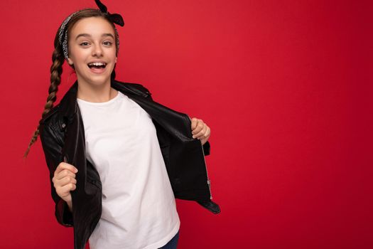 Photo of beautiful happy smiling brunette girl with pigtails wearing stylish black leather jacket and white t-shirt for mockup isolated over red background looking at camera. Empty space