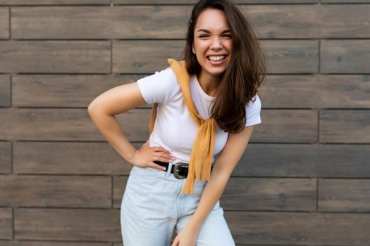 Portrait of successful smiling joyful happy young brunet woman wearing casual white t-shirt and jeans with yellow sweater poising near brown wall in the street and having fun.