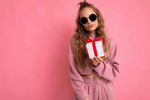 Shot of attractive serious calm young blond curly woman isolated over pink background wall wearing pink sport clothes and modern sunglasses holding gift box looking at camera. Empty space