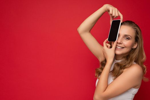 Photo of beautiful smiling young woman good looking wearing casual stylish outfit standing isolated on background with copy space holding smartphone showing phone in hand with empty screen display for mockup looking at camera.