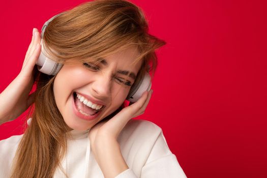 Portrait of charming emotional positive young blonde woman wearing white hoodie isolated over colourful background wearing white wireless bluetooth headphones listening to good music and having fun.