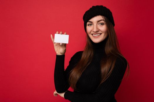 Photo of beautiful smiling young brunette woman wearing black sweater and hat isolated on red background holding credit card looking at camera. copy space