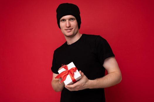 Photo shot of handsome happy young man isolated over red background wall wearing black hat and black t-shirt holding white gift box with red ribbon and looking at camera.