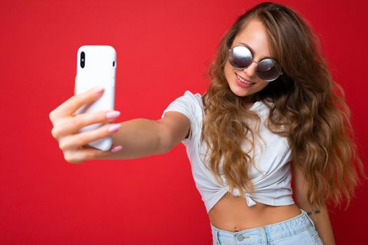 Attractive charming young smiling happy woman holding and using mobile phone taking selfie wearing stylish clothes isolated over wall background.