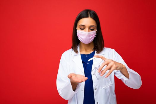 Doctors advice and health protection during coronavirus epidemic. Young woman doctor in protective mask, white coat shows antiseptic in her hands, isolated on background, free space, studio shot.