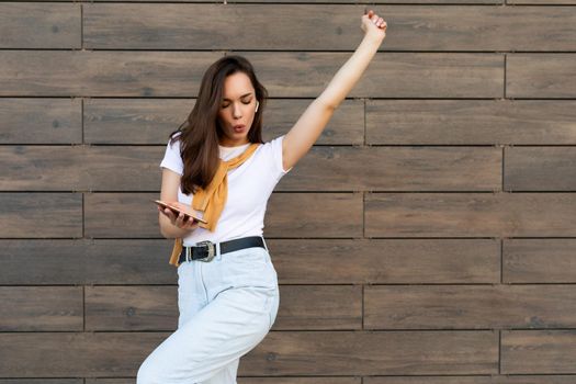 Beautiful brunet young happy woman wearing casual clothes and listening to music via wireless earphones standing in the street holding and using mobile phone and dancing.Copy space