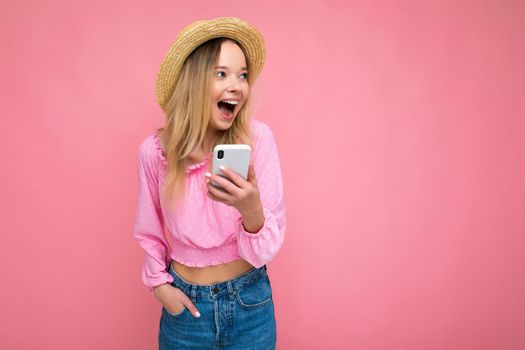 Attractive amazed young blonde woman smiling and having fun standing isolated over pink wall keeping hand in pocket wearing pink blouse and summer hat using mobile phone looking to the side.