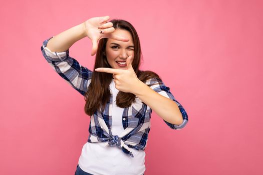 Photo shot of young positive delightful smiling pretty brunette woman with sincere emotions wearing hipster check shirt making frame with hands, taking picture with imaginary camera and standing isolated on pink background with copy space.