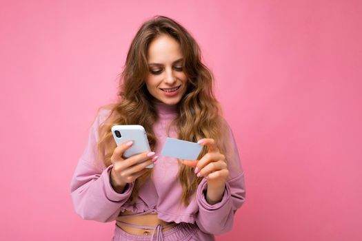 Attractive happy joyful young blonde curly woman wearing pink clothes isolated over pink background using mobile phone paying online shopping through credit card looking at smartphone screen.