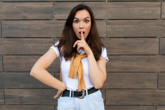 Photo of pretty young brunette woman in trendy outfit. Beautiful attractive female person standing outside in the street and showing shh gesture. Keep secret concept.