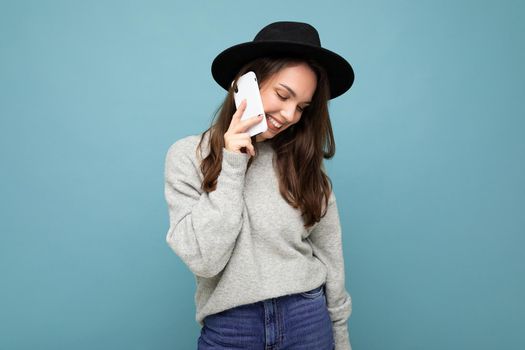 Attractive young shy smiling woman wearing black hat and grey sweater holding smartphone looking down isolated on background.