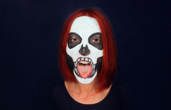 Woman with skull makeup and red hair showing tongue on dark background, Halloween or horror theme