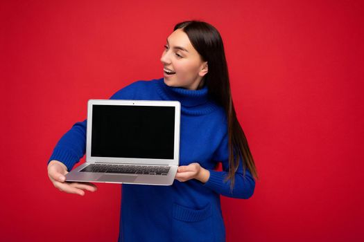 Shot of pretty happy beautiful young brunet woman holding computer laptop looking to the side wearing blue sweater isolated over red wall background. Mock up, free space
