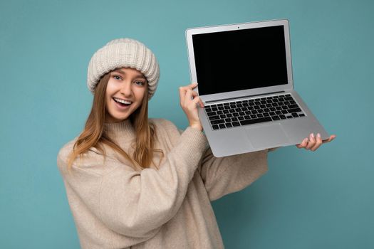 beautiful charming fascinating pretty smiling happy young dark blond feemale student woman in winter warm knitted beige hat holding computer laptop looking at camera wearing beige winter sweater isolated over light blue wall background in studio. Cutout, free empty space for advertising and advertisement