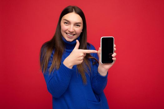 Photo of beautiful smiling young woman good looking wearing casual stylish outfit standing isolated on background with copy space holding smartphone showing phone in hand with empty screen display for mockup pointing at gadjet looking at camera.