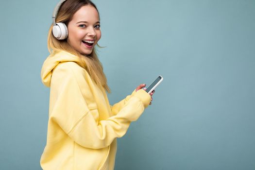 Side-ptofile closeup photo of beautiful smiling young woman wearing stylish casual outfit isolated over colorful background wall wearing white wireless headphones and listening to music and using mobile phone messaging sms looking at camera. copy space