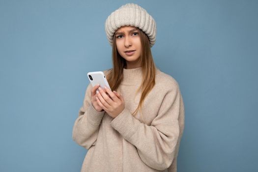 Photo shot of attractive upset dissatisfied good looking young woman wearing casual stylish outfit poising isolated on background with empty space holding in hand and using mobile phone messaging sms looking at camera.