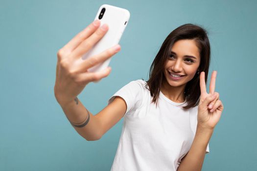 Photo of beautiful smiling happy young brunette woman wearing casual white t-shirt isolated over wall blue background holding and using mobile phone taking selfie looking at gadjet screen and showing peace gesture.