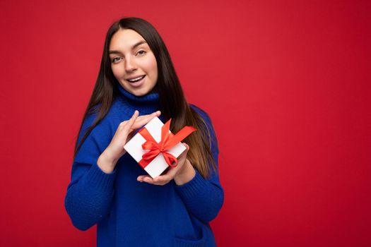 Shot of attractive happy smiling young brunette woman isolated over red background wall wearing blue casual sweater holding white gift box and looking at camera. Empty space