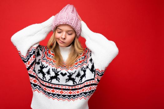 Photo of beautiful thoughtful sad young blonde woman isolated over red background wall wearing winter sweater and pink hat looking to the side and thinking.