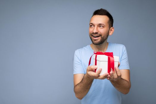 Photo shot of handsome positive brunette unshaven young man isolated over blue background wall wearing blue t-shirt holding white gift box with red ribbon and looking to the side. Copy space, mockup