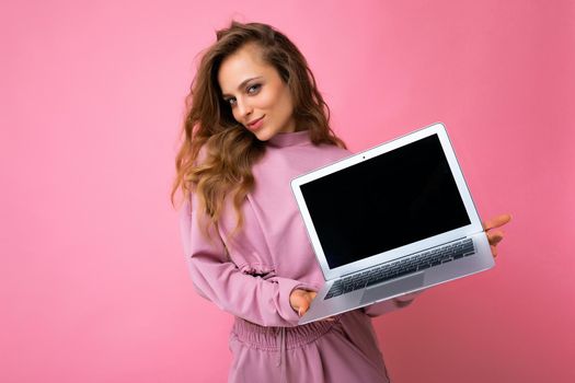 Photo of beautiful blond curly young woman lookind at camera holding computer laptop with empty monitor screen with mock up and copy space wearing pink hoodie isolated over pink wall background.