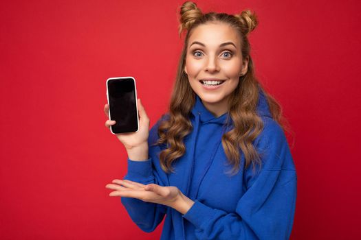 Attractive smiling young blonde woman wearing stylish blue hoodie isolated on red background with copy space holding smartphone showing phone in hand with empty screen display pointing at gadjet looking at camera.