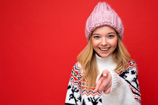 Photo of young positive happy smiling beautiful woman with sincere emotions wearing stylish clothes isolated over background with copy space and pointing at camera.