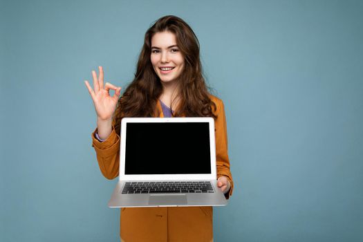 Photo of Beautiful smiling satisfied happy young woman holding computer laptop looking at camera wearing yellow jacket showing okay gesture isolated over blue wall background. Empty free space, mock up