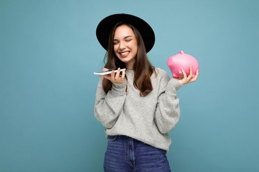 Photo of happy positive smiling laughing young beautiful brunette woman wearing stylish sweater and black hat isolated over blue background with empty space, holds pink piggy bank and uses mobile phone communicating online.