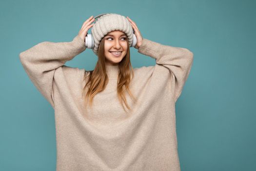 Beautiful happy smiling young blonde woman wearing beige winter sweater and hat isolated over blue background wearing white wireless bluetooth headphones listening to cool music enjoying and looking to the side. copy space