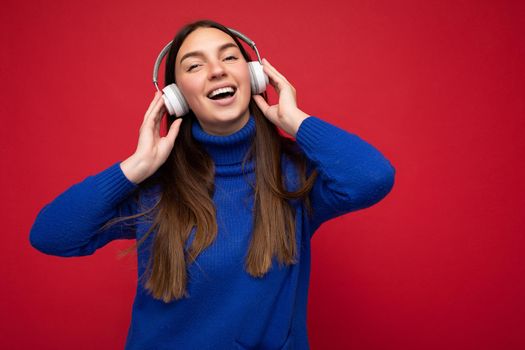 Attractive positive young brunette woman wearing blue sweater isolated over red background wall wearing white bluetooth earphones listening to cool music and having fun.