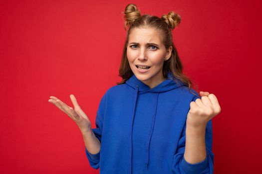 Photo of emotional agressive young beautiful blonde woman with sincere emotions wearing blue pullover isolated over red background with free space and showing fist.