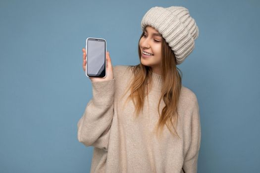 Photo of beautiful smiling young woman good looking wearing casual stylish outfit standing isolated on background with copy space holding smartphone showing phone in hand with empty screen display for mockup looking to the side.