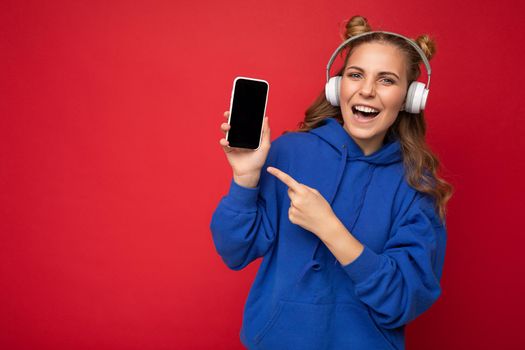 Photo of beautiful smiling young woman wearing stylish blue hoodie isolated over red background wearing white bluetooth wireless headphones and listening to music and showing mobile phone with empty space for mockup and pointing finger at gadjet. copy space