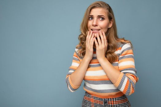 Portrait of young beautiful blonde curly woman with sincere emotions wearing stylish striped sweater isolated on blue background with empty space and looking stressed and nervous.